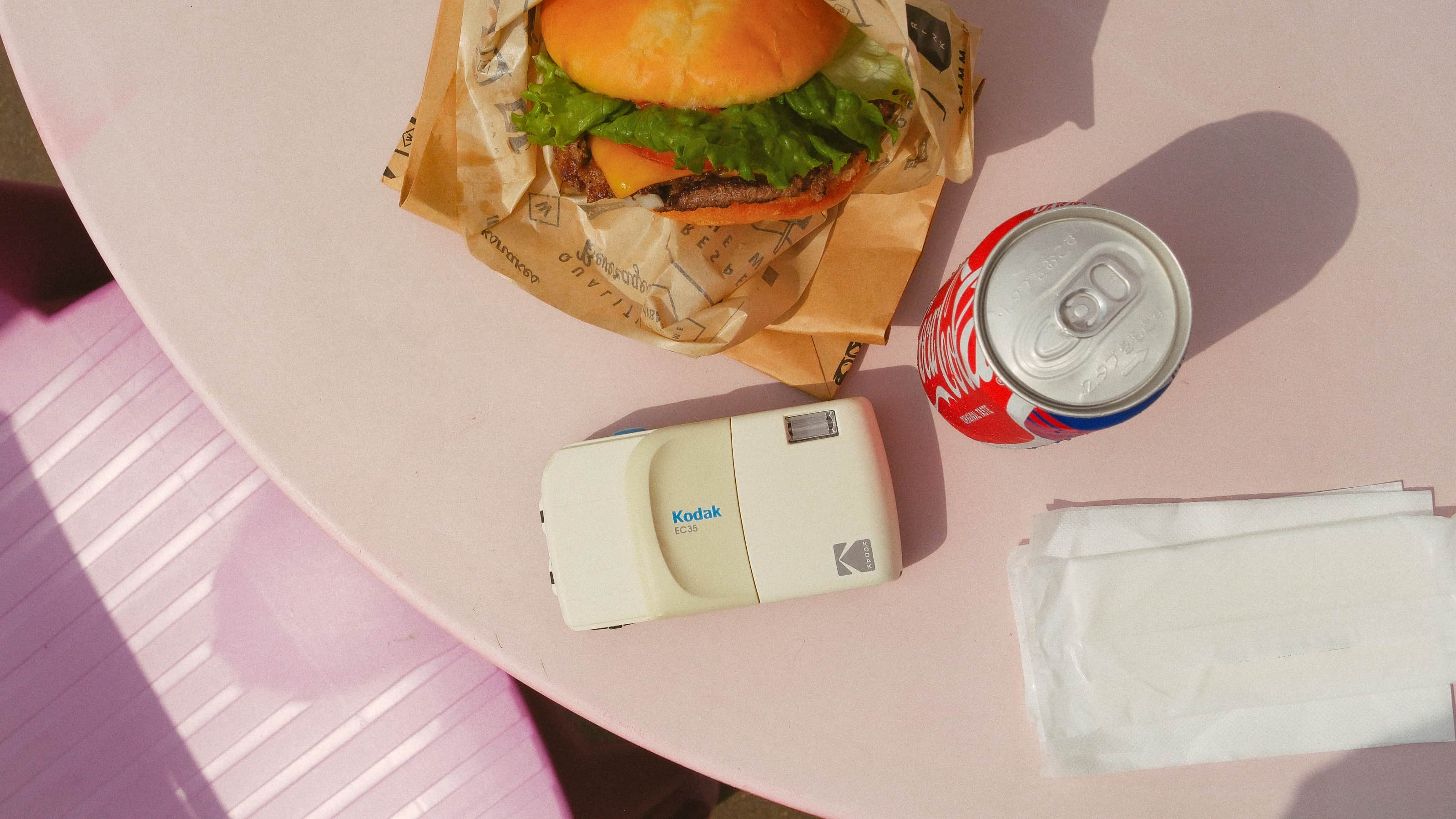 Photograph of the white Kodak EC35 film camera on a table beside a burger and a can of Coke