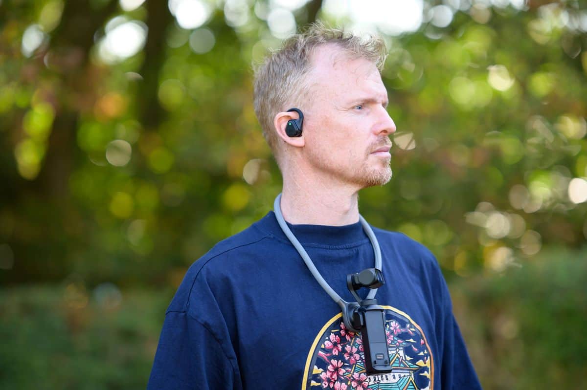 Tim Coleman wearing the Insta360 POV Head Tracker accessory in his ear, with the Luna Ultra camera on his chest attached to a neck mount, outdoors with dappled light in the background foliage
