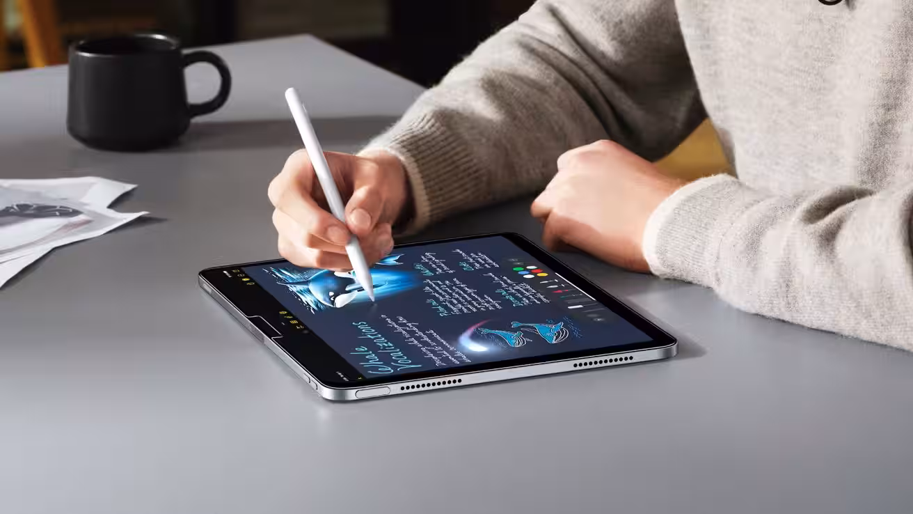 Person in a light sweater using a white stylus to draw colorful digital artwork and handwritten notes on a tablet lying flat on a gray desk with a black mug nearby