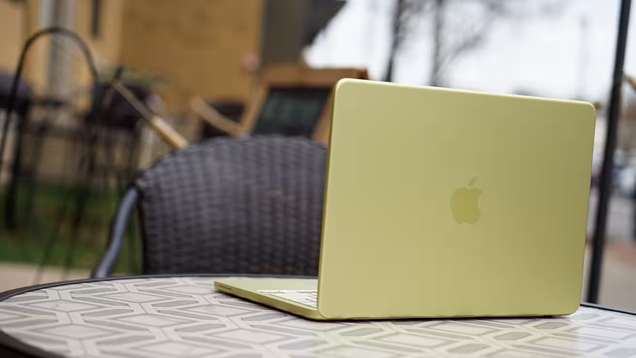 Light green Apple laptop open on a patterned outdoor cafe table, with a dark wicker chair and blurred buildings and trees in the background