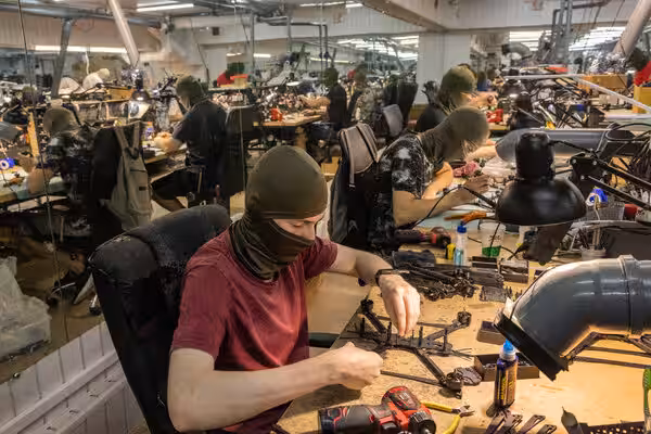 Several people in olive-green balaclavas assemble components at individual workbenches. They work in a brightly lit, busy workshop.